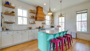 A kitchen with white cabinets, a turquoise island, wooden shelves, and pink bar stools.