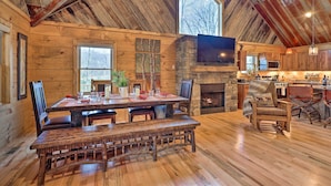 L’intérieur d’un chalet rustique est agrémenté d’une table à manger en bois, d’une cheminée en pierre et d’un grand téléviseur fixé au mur.
