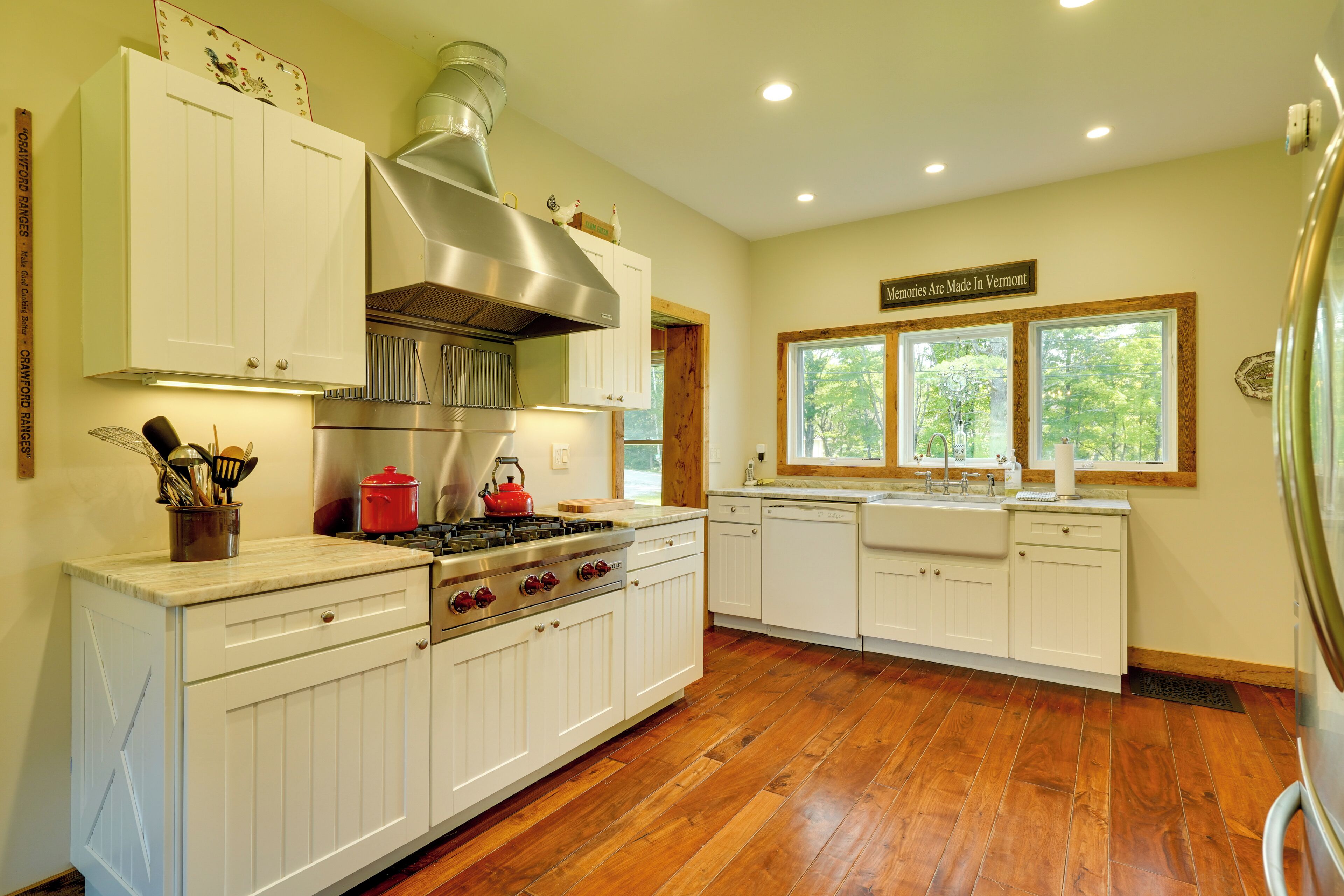 A kitchen with white cabinetry, a stainless steel range hood, a red kettle, and a window with a view of trees.