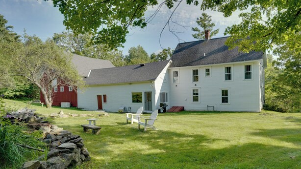 A two-story house with a red barn, a stone wall, and a grassy yard with a picnic table and chairs.