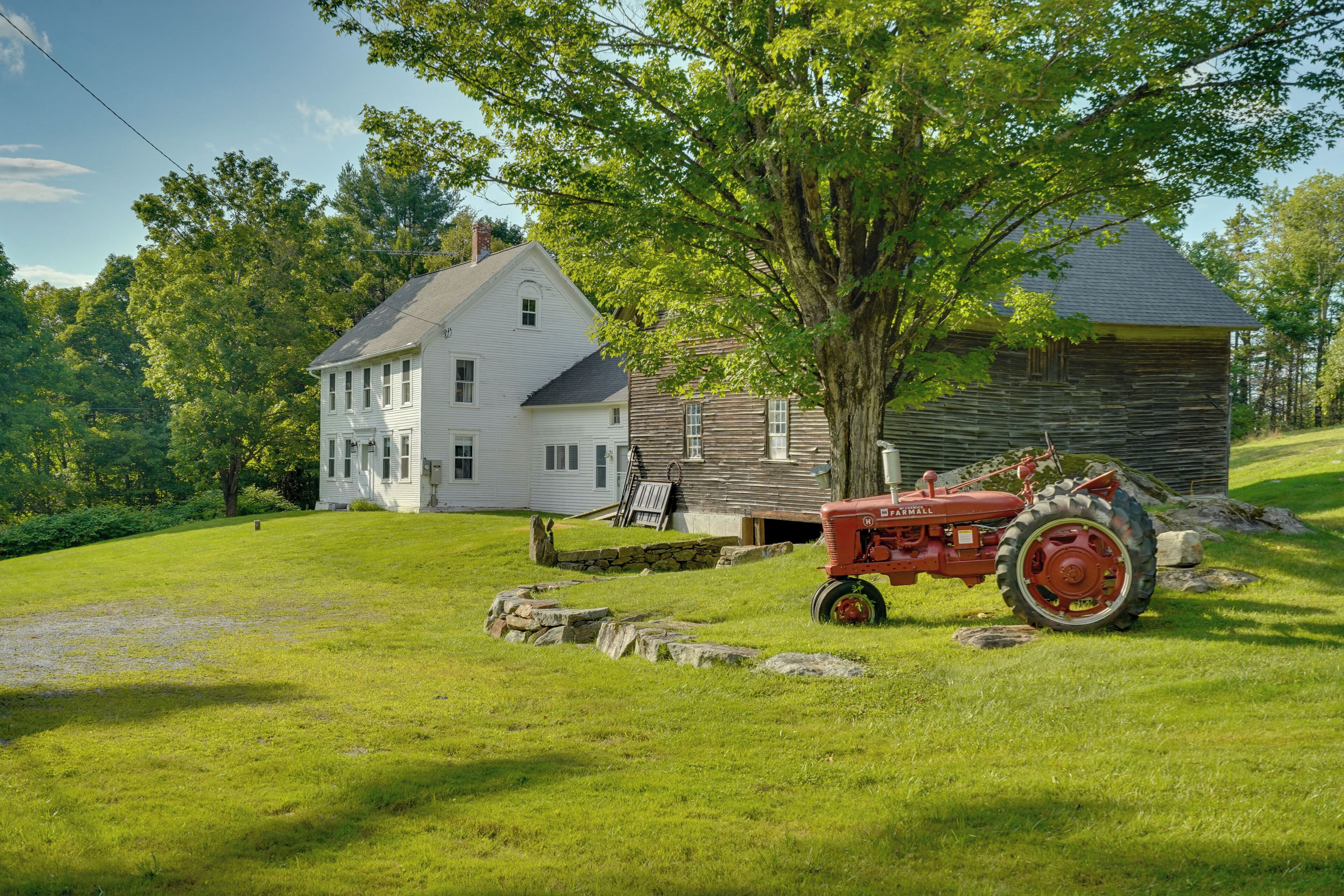 A two-story house with a stone foundation, a large tree in front, and a red tractor parked on the grass.