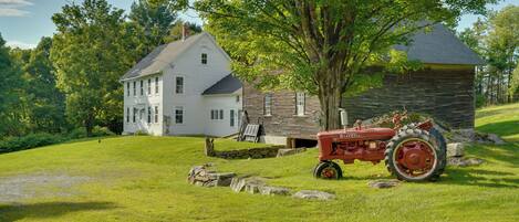 A two-story house with a stone foundation, a large tree in front, and a red tractor parked on the grass.