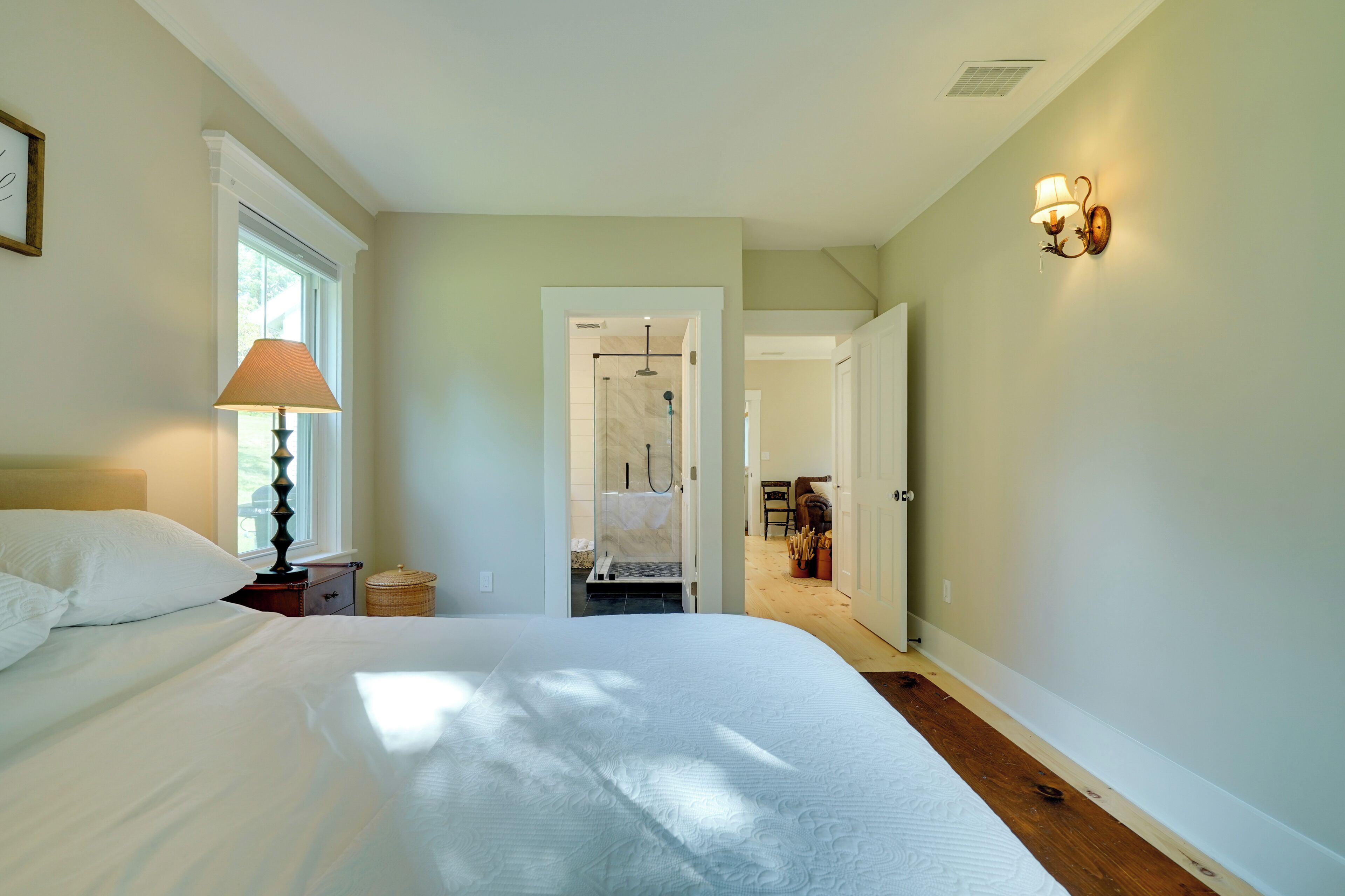 A bedroom with a large bed, a nightstand, a lamp, and a view of a bathroom with a shower.
