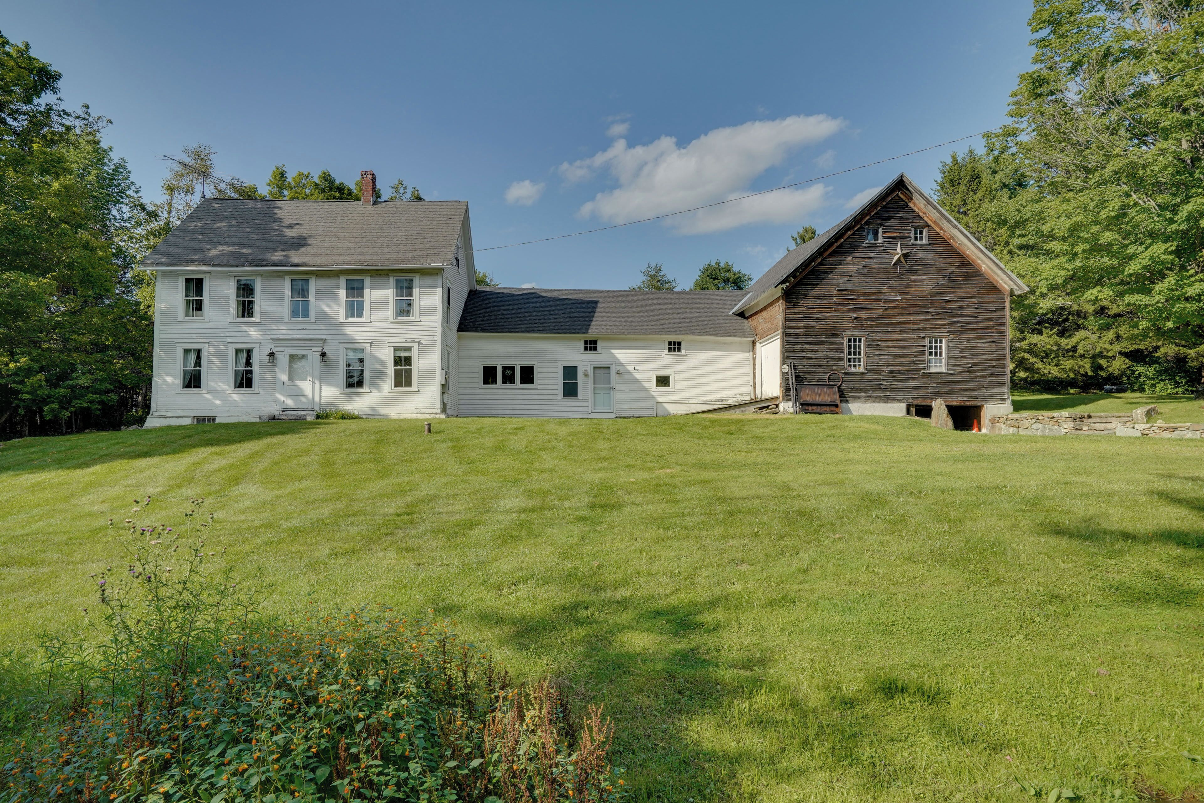 Une grande maison blanche avec un toit sombre et une grange en bois ornée d’une étoile sur le côté.