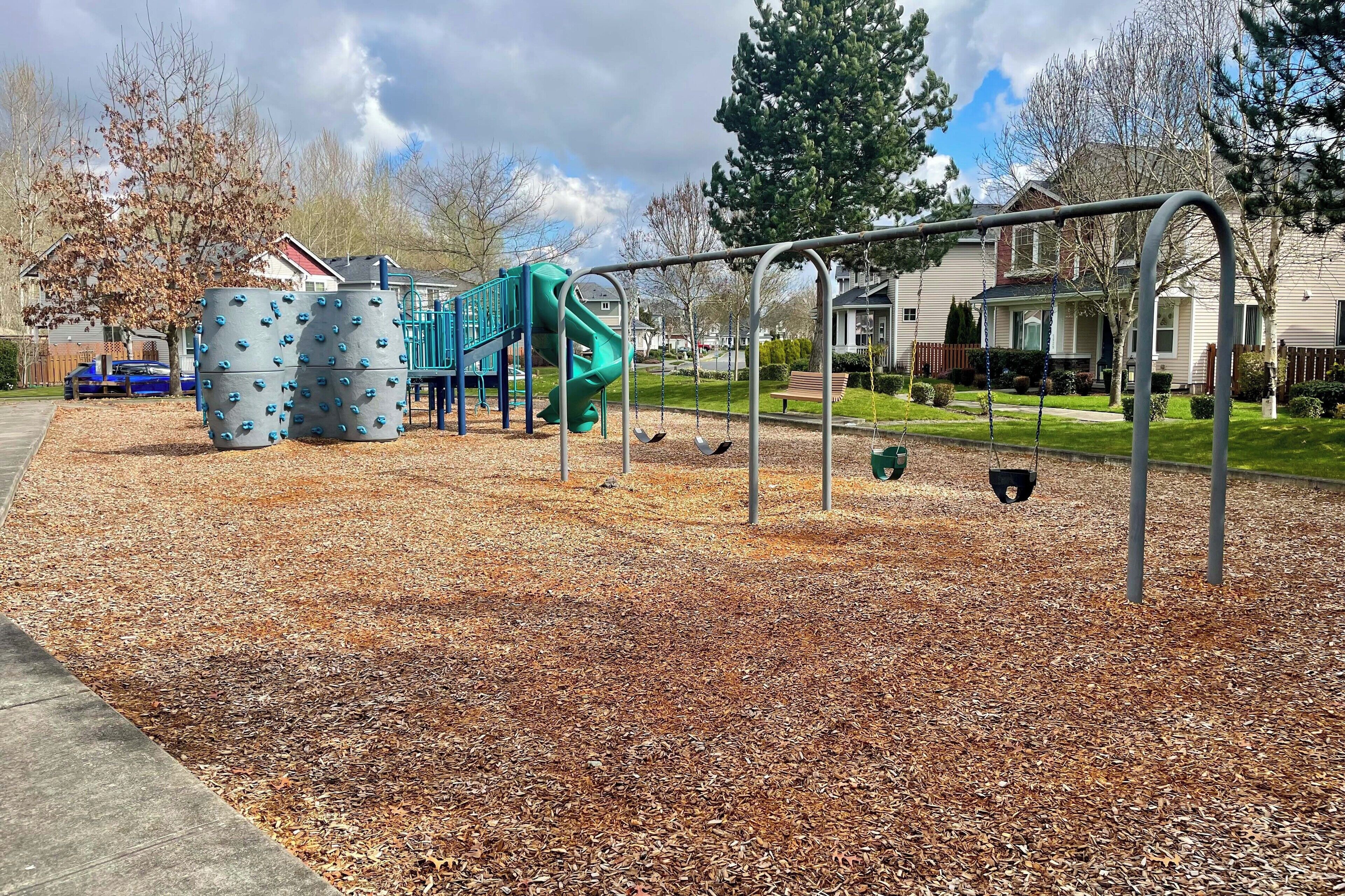 A playground with a slide and climbing structures, surrounded by a grassy area and residential houses.