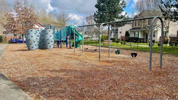 A playground with a slide and climbing structures, surrounded by a grassy area and residential houses.