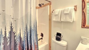 A bathroom with a shower curtain depicting a forest scene, a white bathtub, a toilet with a black bag on it, and a white sink. - Murphy Family Cabin w/ Fire Pit & Forest Views! (Murphy)