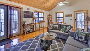 A cozy living room with a wooden ceiling, a glass coffee table, a sofa with decorative pillows, and a TV mounted on the wall. - Murphy Family Cabin w/ Fire Pit & Forest Views! (Murphy)