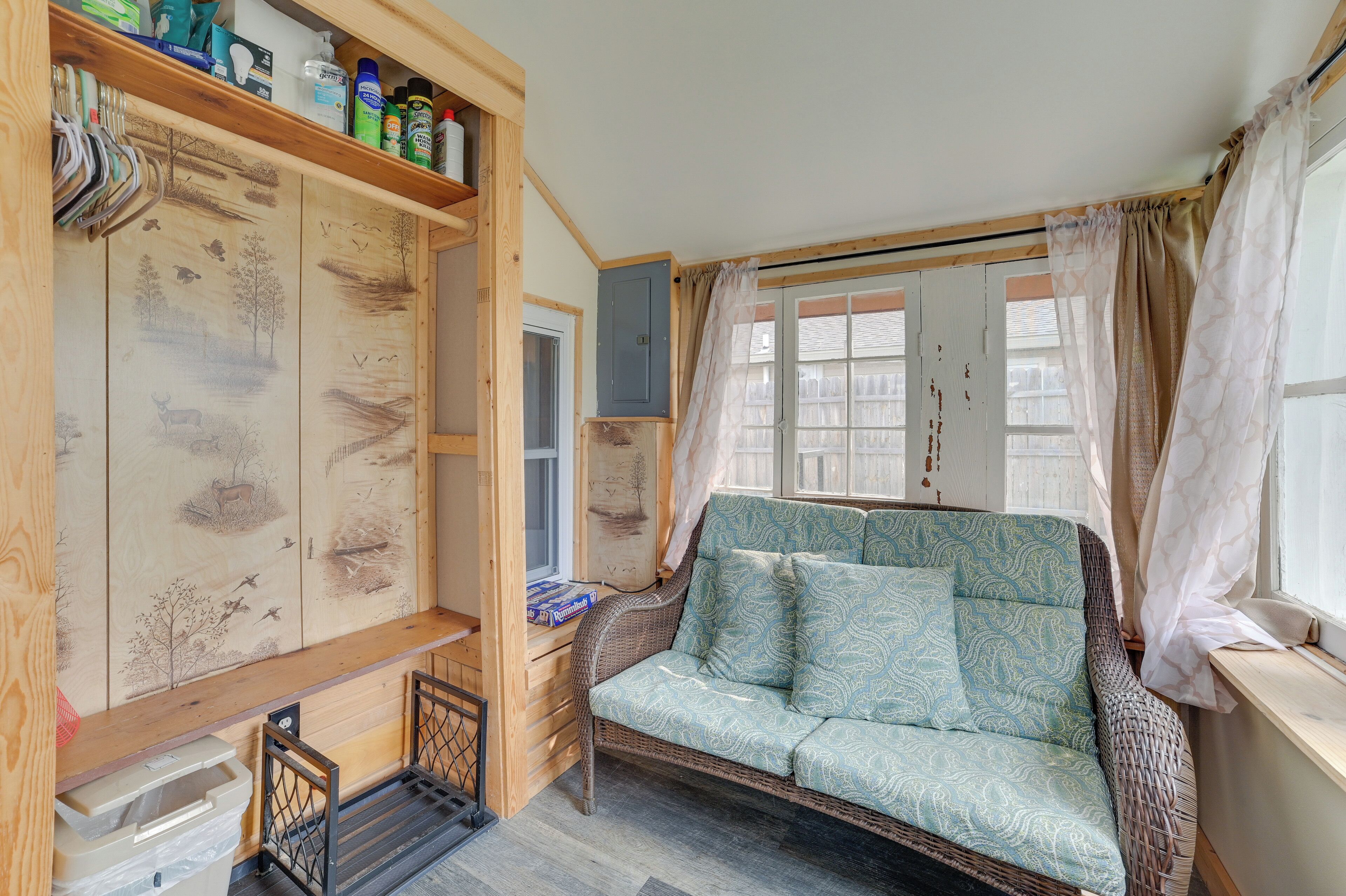 Una habitación acogedora con un sofá de mimbre, estanterías de madera con diversos objetos, y vistas a través de una ventana con cortinas.