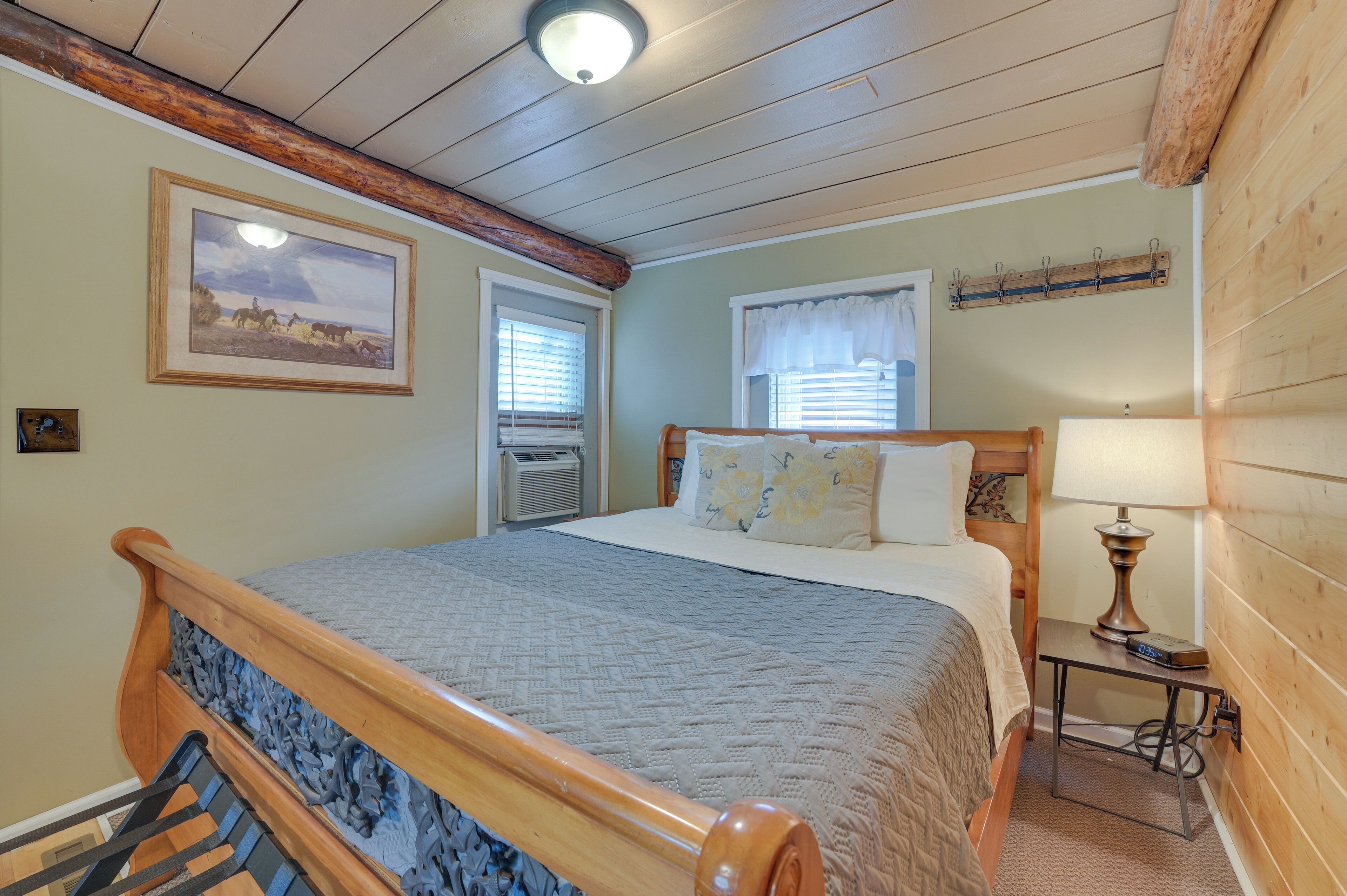 A bedroom with a wooden bed, a nightstand with a lamp, a framed picture on the wall, and a window with blinds.