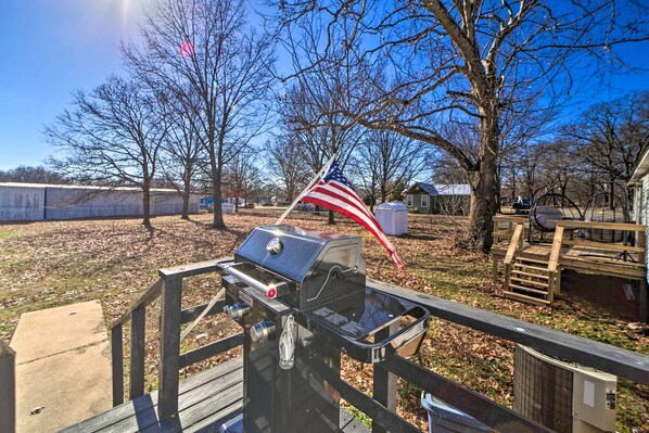 A barbecue grill with an American flag, a wooden deck, and a fenced yard with leaf-covered ground.
