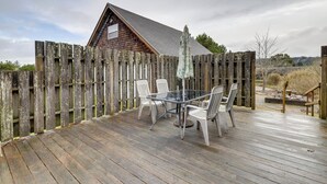 A wooden deck with a glass table and chairs, a wooden fence, and a building with a sloped roof in the background.
