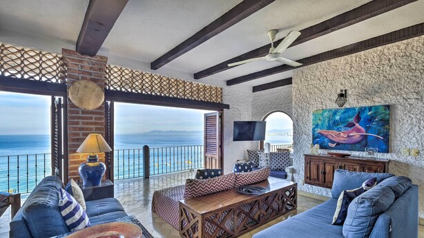 A spacious living area with a large sofa, a coffee table, and a TV. The room has a view of the ocean and features a stone wall and wooden beams. - Mid-century Modern Condo w/ Patio & Ocean Views! (Puerto Vallarta)