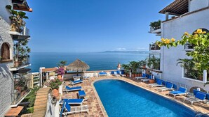 A swimming pool with blue lounge chairs, a view of the sea, and a building with balconies and potted plants. - Mid-century Modern Condo w/ Patio & Ocean Views! (Puerto Vallarta)