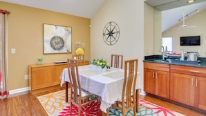 Uma sala de jantar com uma mesa posta para quatro pessoas, cadeiras de madeira, um objeto decorativo na parede e uma cozinha com armários de madeira e bancada preta.