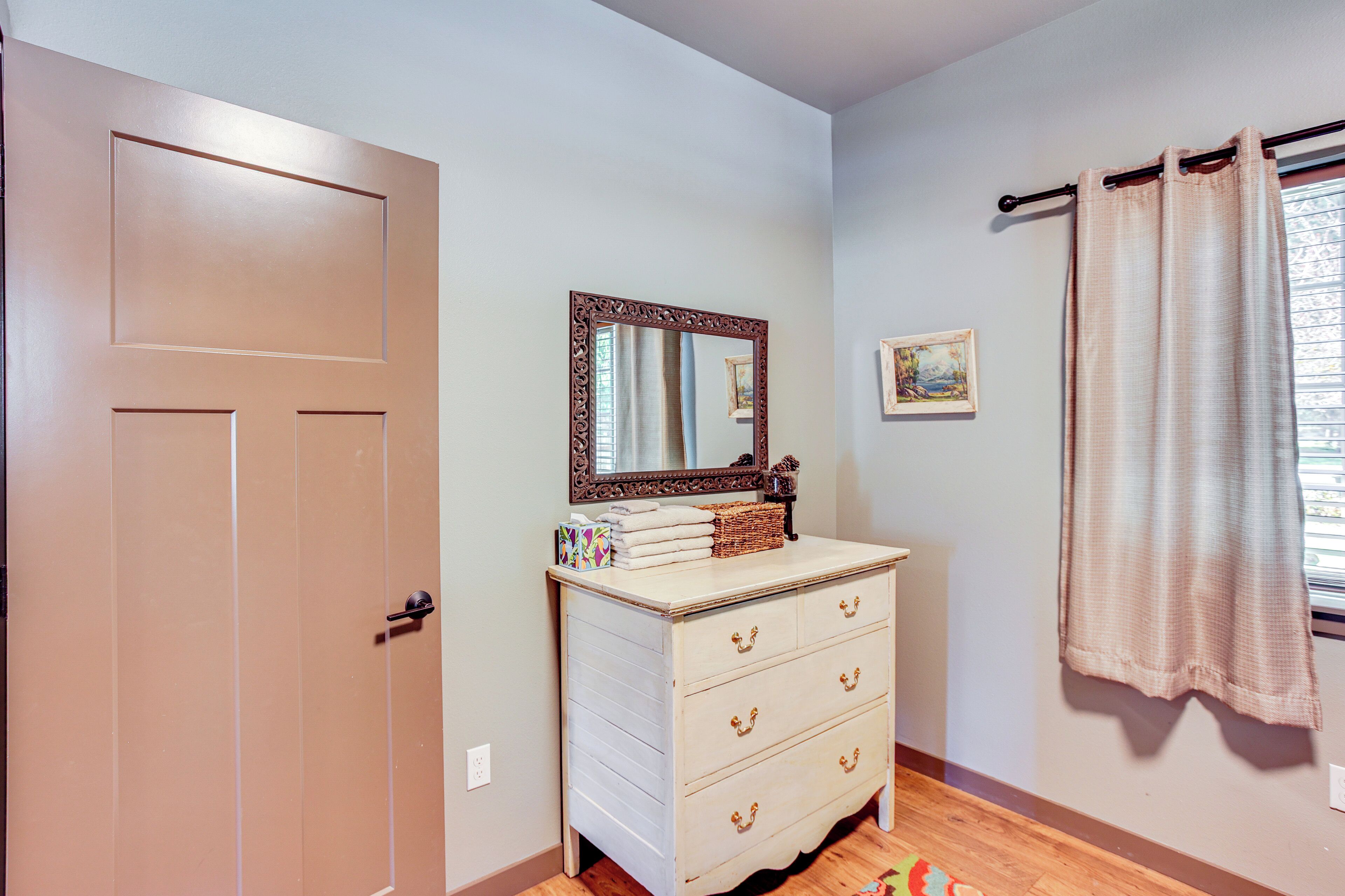 A room with a wooden door, a dresser with drawers, a mirror, a framed picture, and a window with curtains.
