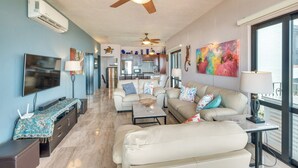 A spacious living room with a large TV, comfortable seating, a ceiling fan, and a view to the kitchen.