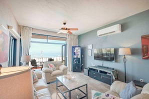 Sala de estar com sofá, mesa de centro e televisão. Varanda com vista para o oceano.