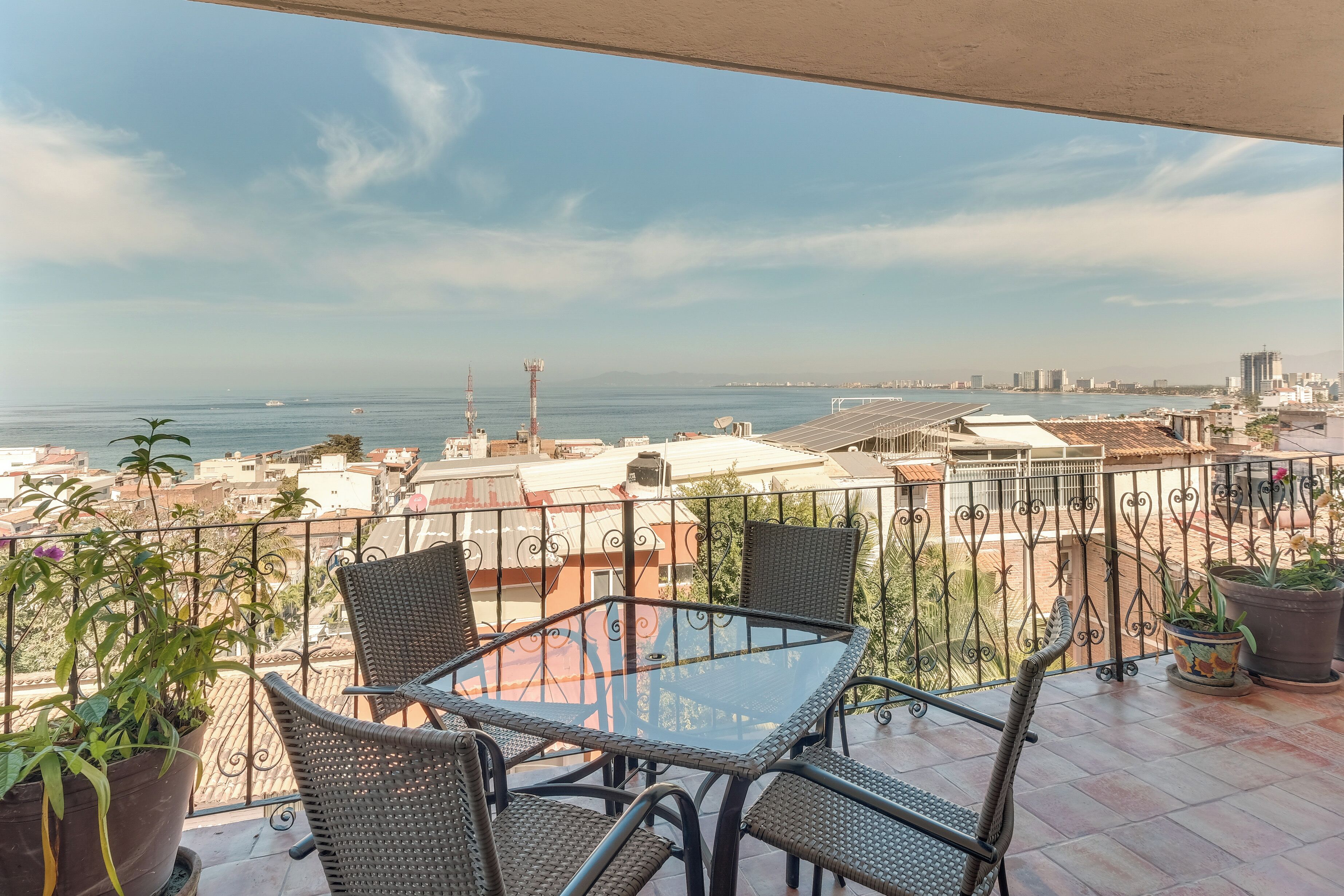 A balcony with a glass table and wicker chairs, overlooking a cityscape and the sea.