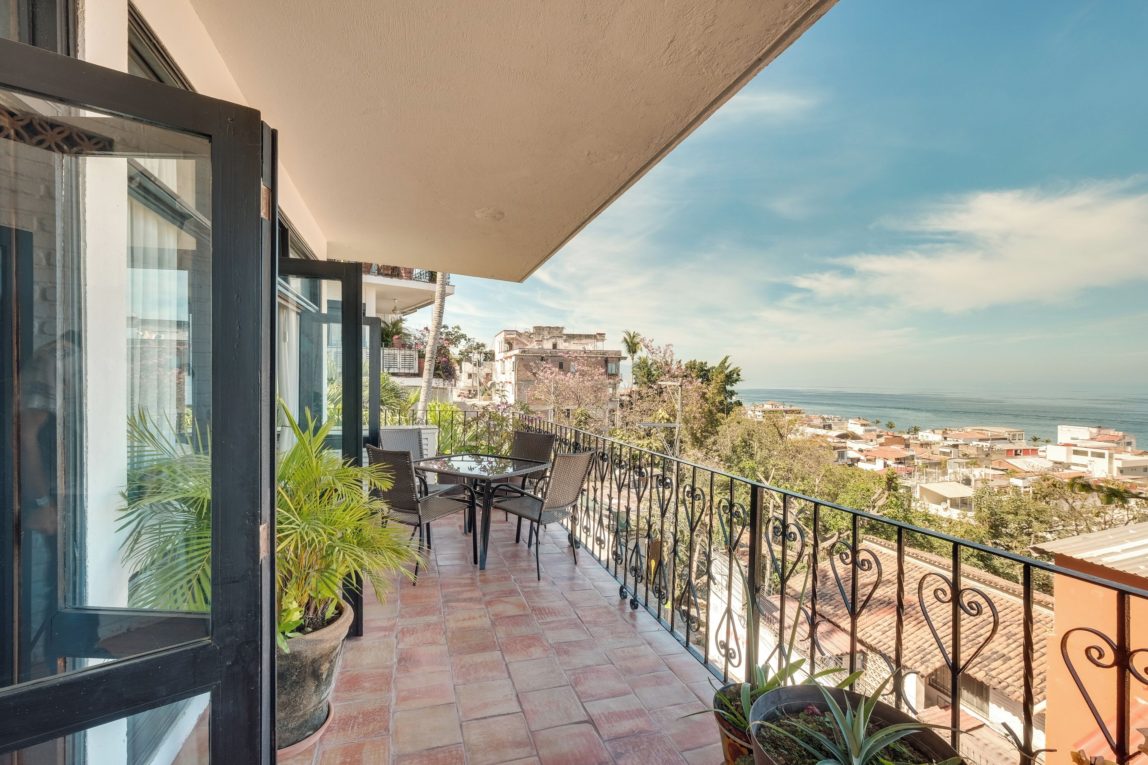 A balcony with a table and chairs, potted plants, and a view of a coastal town.
