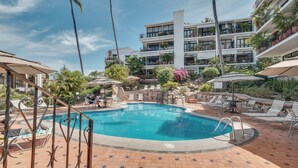 A pool area with lounge chairs and umbrellas, surrounded by a multi-story building with balconies and plants.