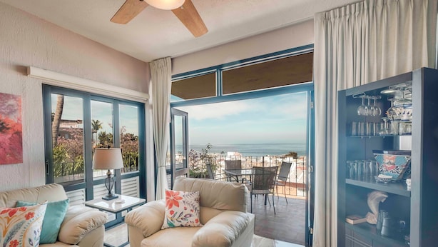 A living room with a sofa, armchairs, a coffee table, and a cabinet. There is a large window with a view of the ocean and a balcony with outdoor furniture.