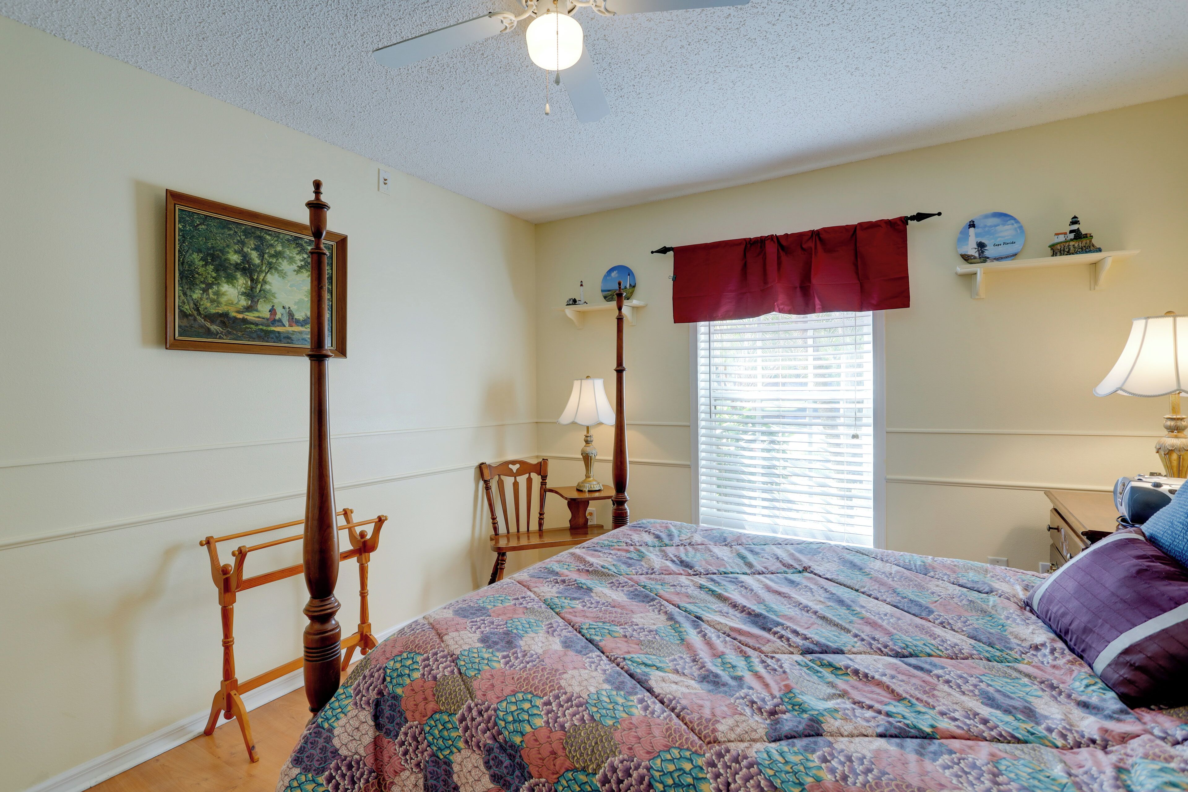 A bedroom with a bed, a wooden chair, a standing lamp, a window with blinds, and a wall-mounted shelf with decorative items.
