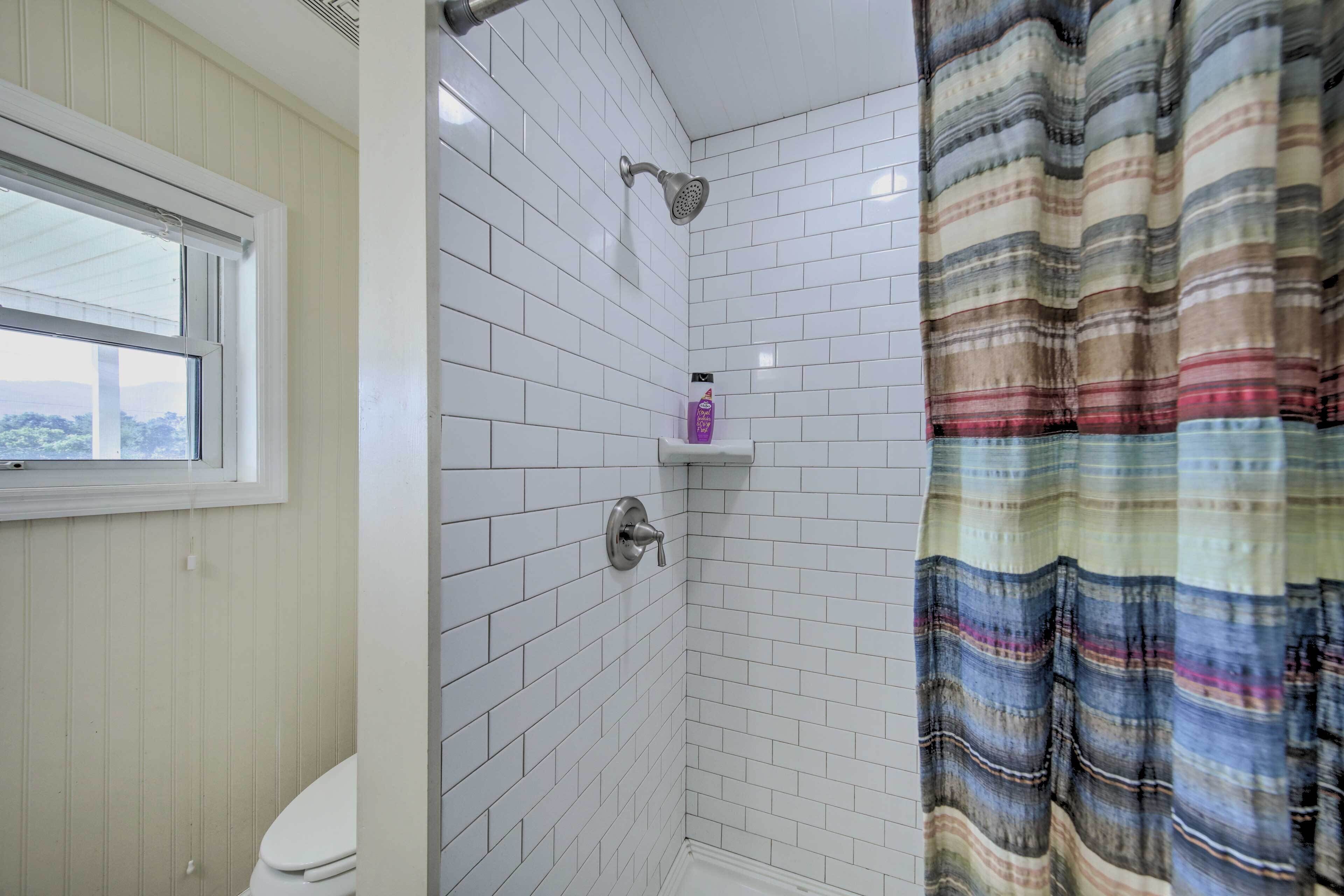 A bathroom with white tiled walls, a shower with a handheld showerhead, a window, and a striped shower curtain.