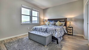 Quarto com cama grande, um banquinho, mesa de cabeceira e janela.