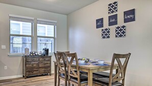 Uma sala de jantar com mesa e cadeiras de madeira, uma janela com persianas, um aparador de madeira e decoração na parede com texto.