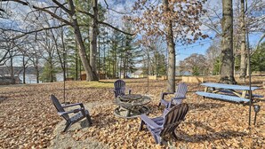 A park with a fire pit, chairs, and picnic tables surrounded by trees and a lake. - Fire Pit & Grill: Lakeview Mecosta Cottage! (Mecosta)