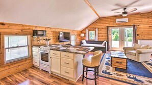 A cozy cabin with a kitchen area featuring a stove, microwave, and a dining table with chairs. There is a ceiling fan, a TV mounted on the wall, and a large window with curtains. - Quaint Studio w/ Shared Beach: Walk to Suttons Bay (Suttons Bay)