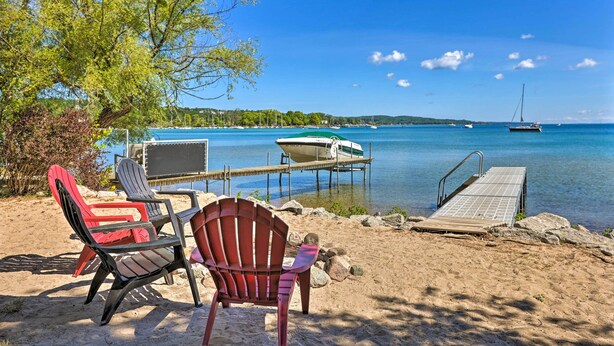 A beach with two Adirondack chairs, a dock extending into calm water, and a sailboat in the distance. - Quaint Studio w/ Shared Beach: Walk to Suttons Bay (Suttons Bay)