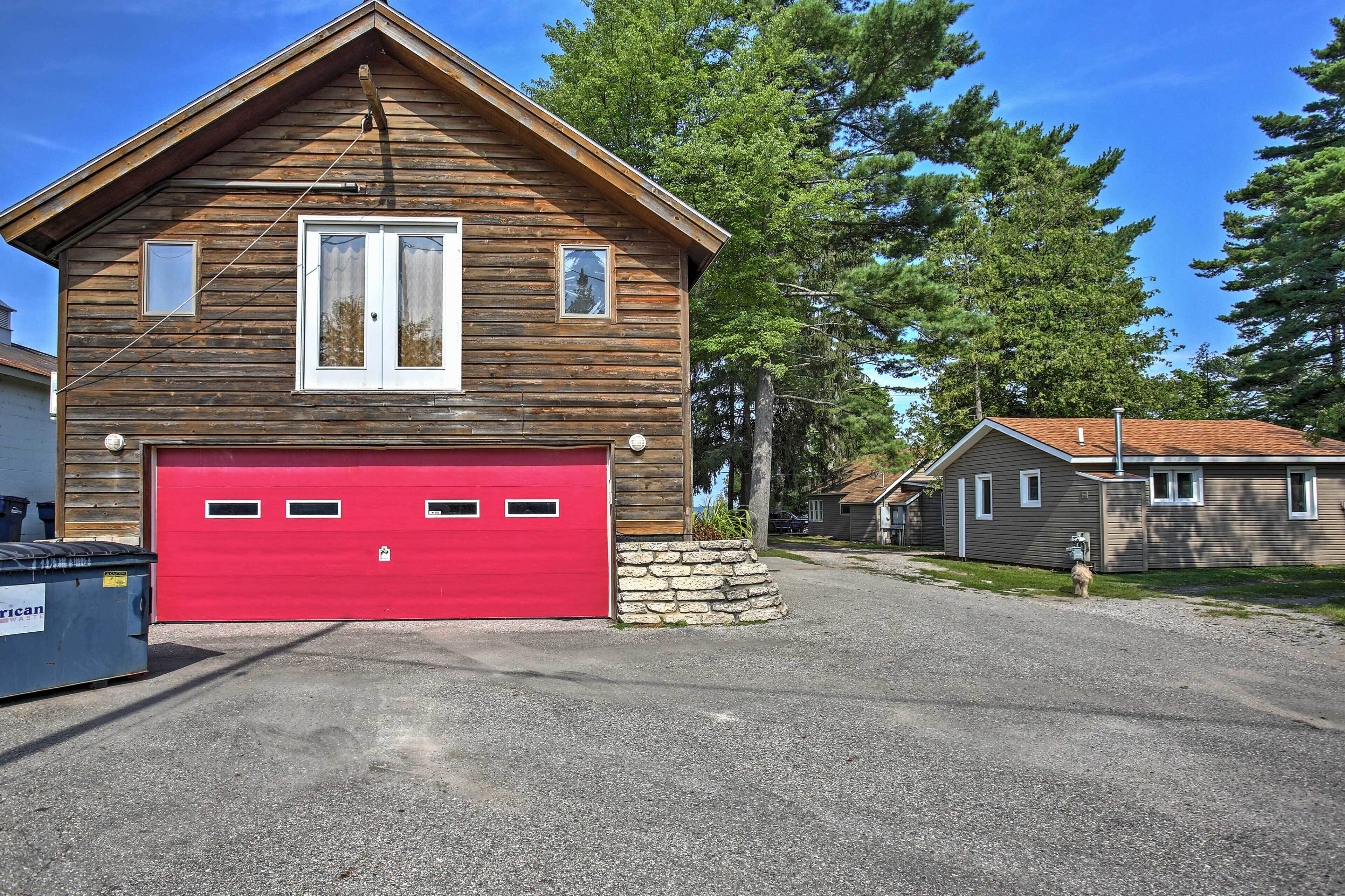 A two-story wooden house with a red garage door, a stone foundation, and a driveway leading to a gravel area.
