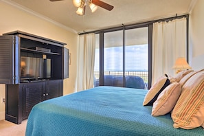 A bedroom with a large bed, a television on a cabinet, a balcony with curtains, and a ceiling fan. - Family Condo: Resort Pool Access & Ocean View (Dauphin Island)
