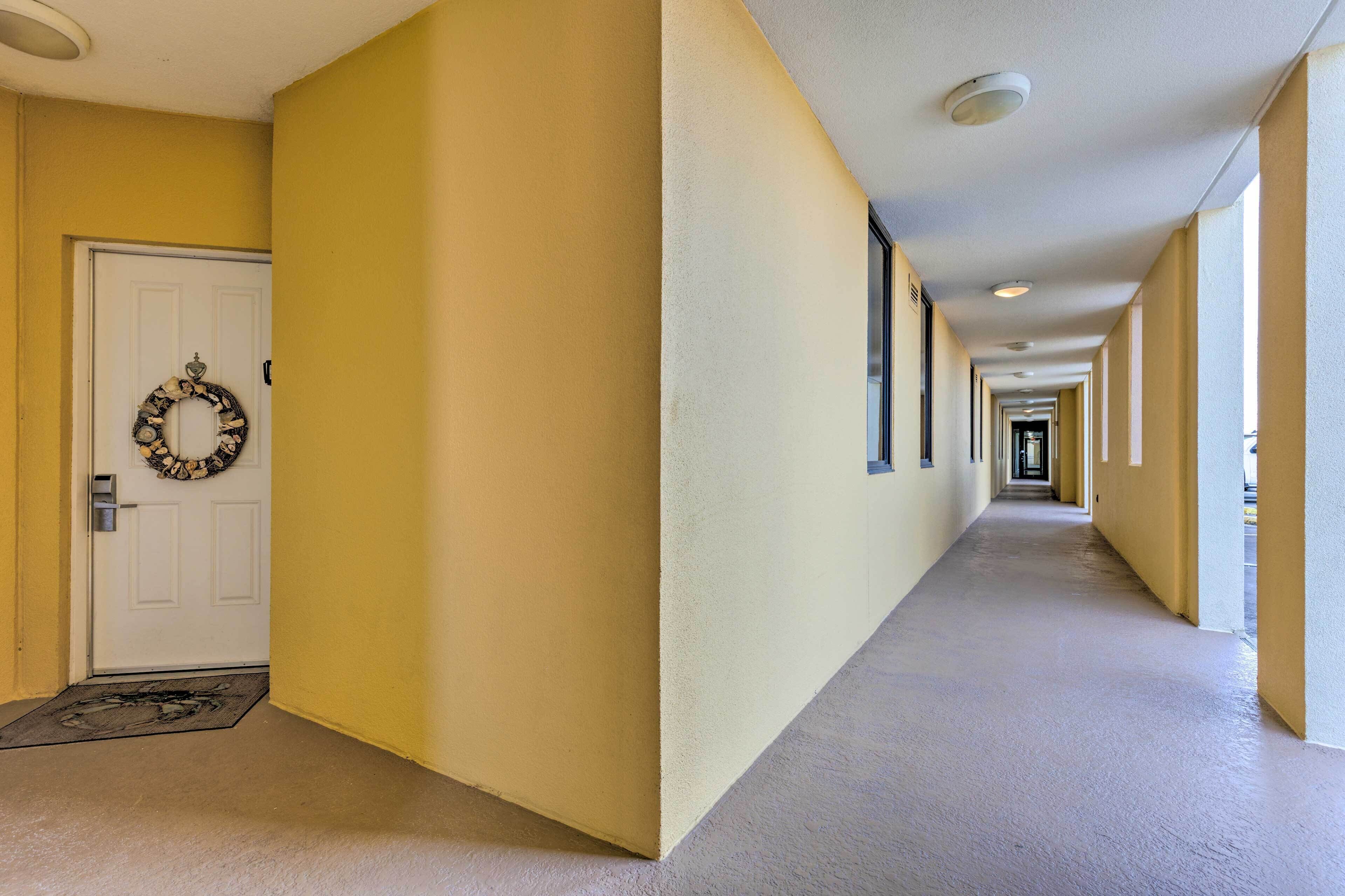 A long, narrow hallway with a white door on the left, a doormat, and a wreath on the door. The walls are painted yellow, and there are windows along the right side.
