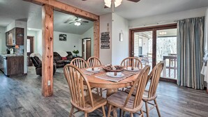 A dining area with a wooden table and chairs, a kitchen in the background, and a large window with curtains.
