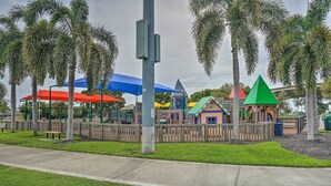 Um parquinho com estruturas coloridas para brincar, rodeado por palmeiras e coberto por guarda-sóis azuis e vermelhos.