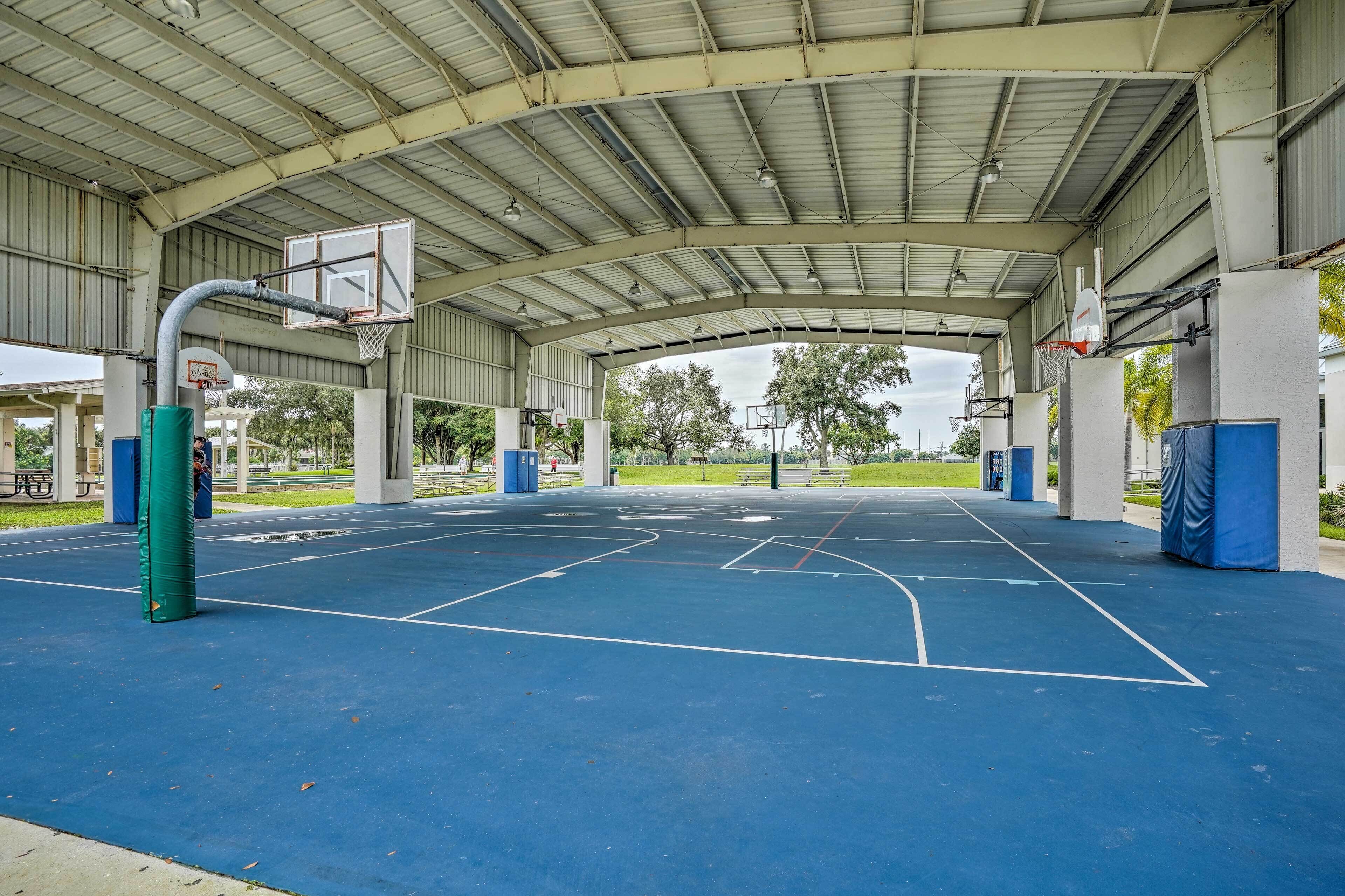 An indoor basketball court with a metal roof, green exercise equipment, and a clear view of the surrounding park.