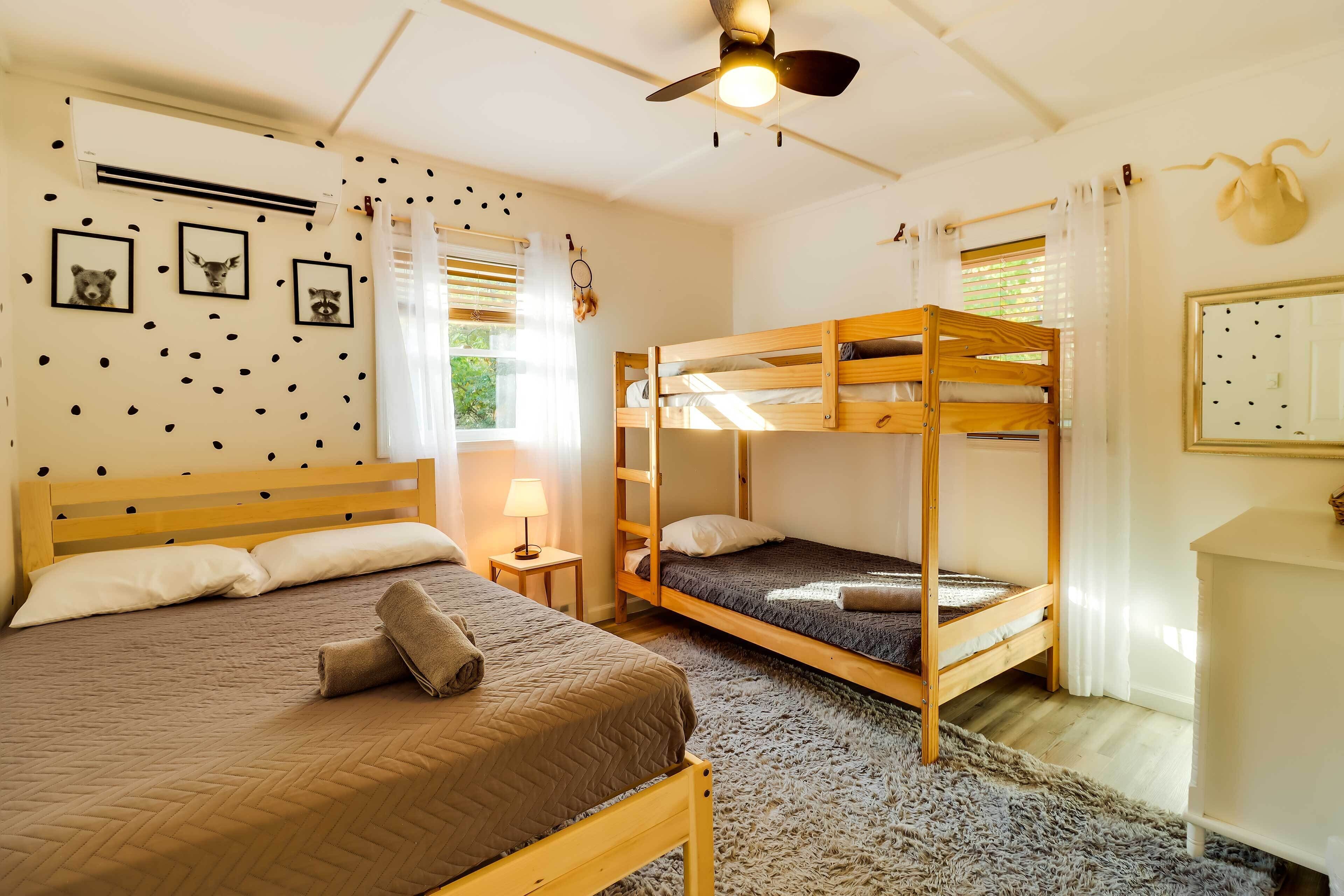 Una habitación con una cama individual, una cama litera, un ventilador de techo, una mesita con una lámpara y aire acondicionado de pared.
