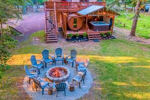 A wooden cabin with a deck, a hot tub, and a fire pit surrounded by Adirondack chairs.