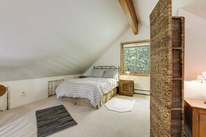 Une chambre avec un plafond en pente, un lit avec une literie à rayures, une petite table de chevet avec une lampe, une armoire en bois et une fenêtre donnant sur des arbres.