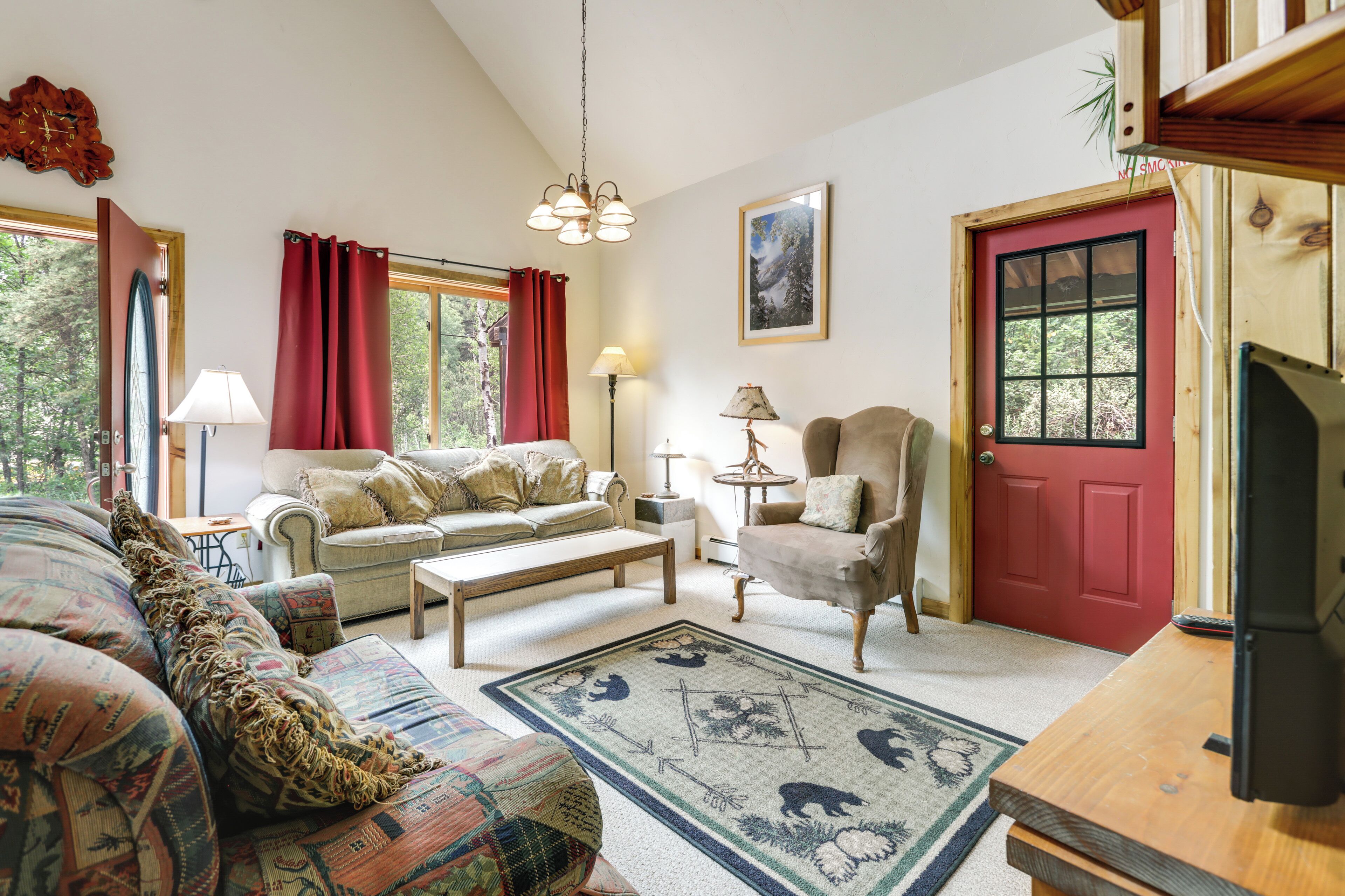 A cozy living room with a sofa, armchair, and coffee table, a red door, and a window with curtains.
