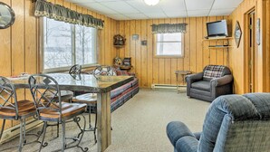 A cozy cabin interior with a wooden dining table, chairs, a sofa, and a television on a shelf. - Tranquil Marenisco Cabin on Lake Gogebic! (Marenisco)