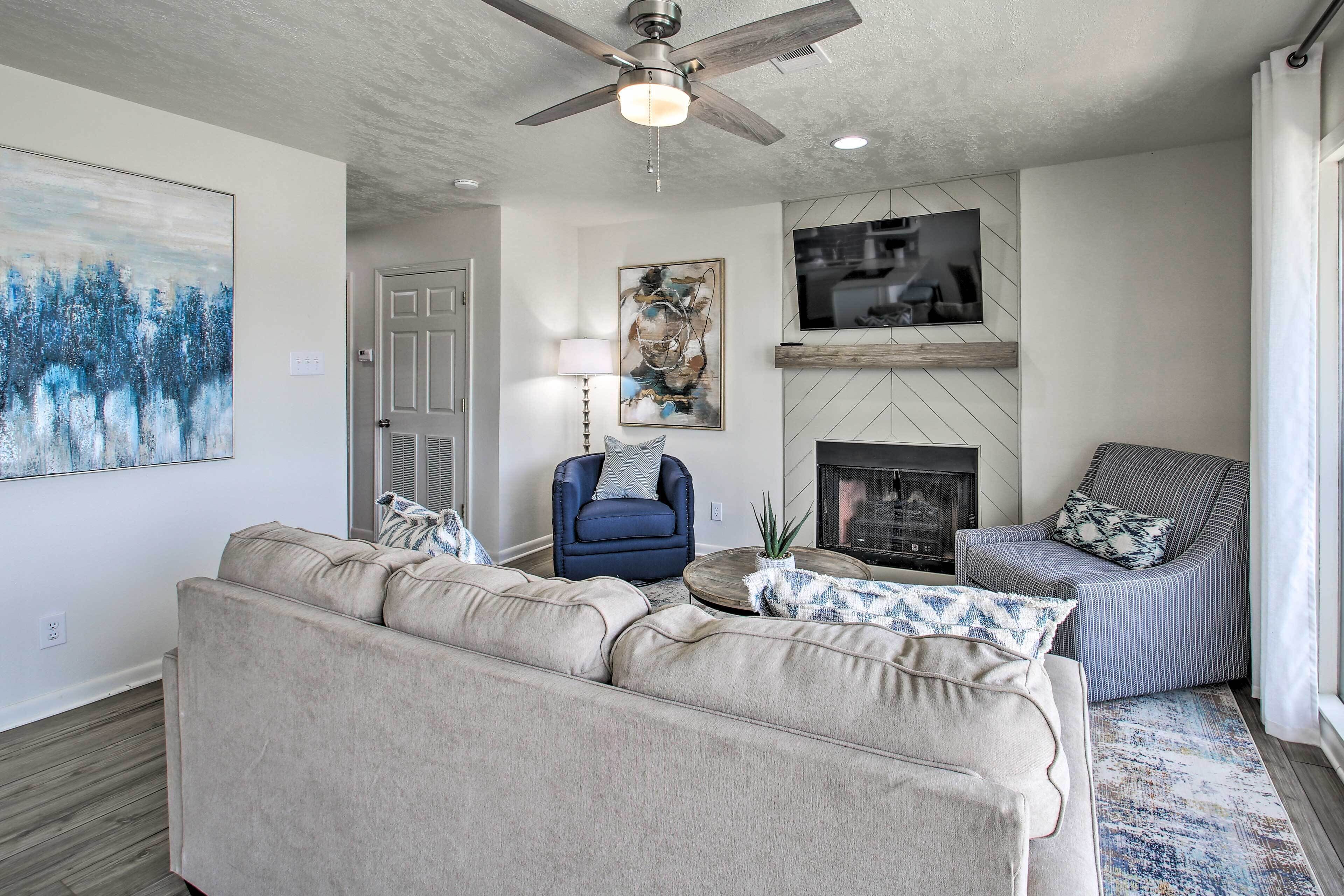 A living room with a beige sofa, a blue armchair, a striped armchair, a fireplace, and a ceiling fan.