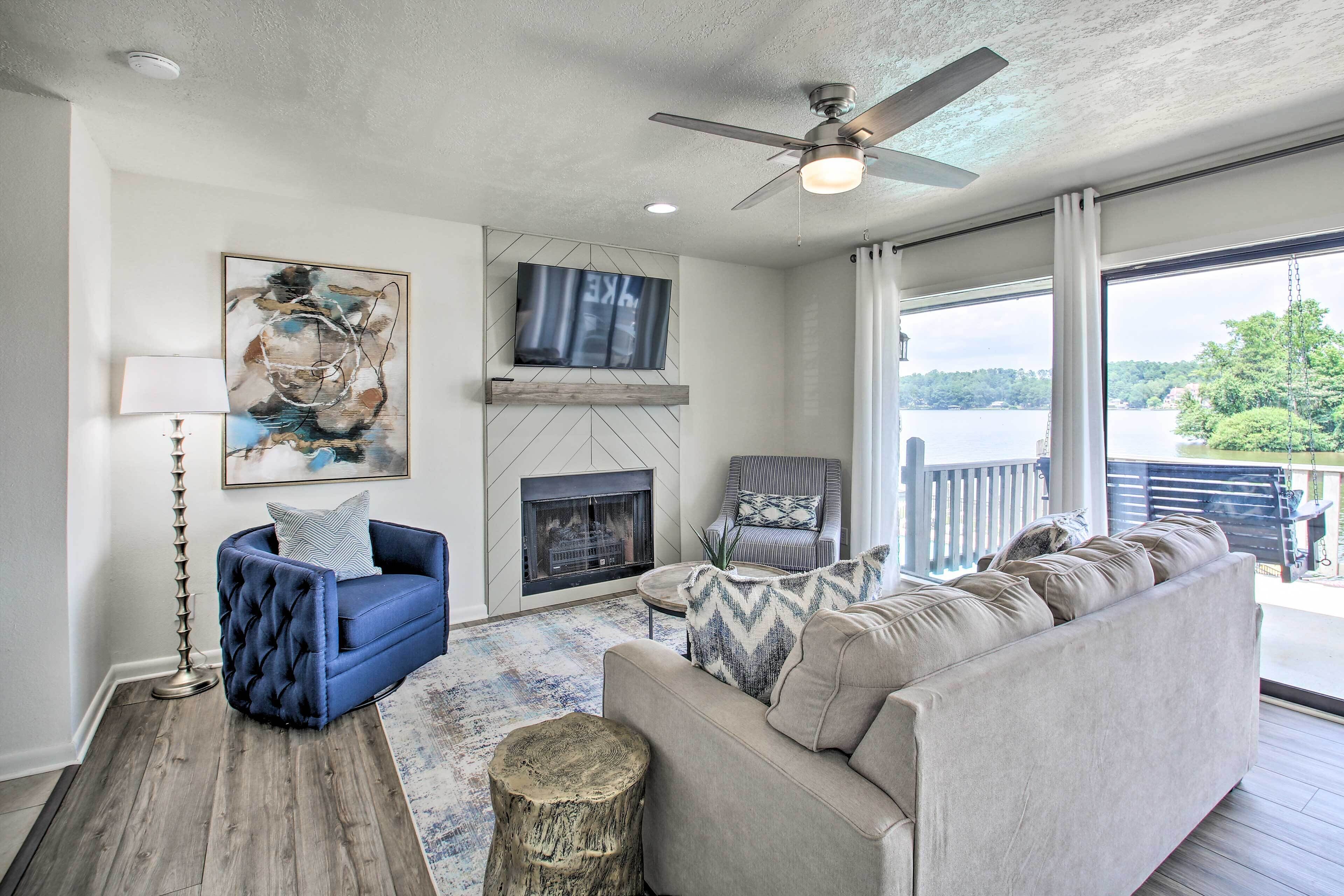 A living room with a fireplace, a ceiling fan, and a large window overlooking a lake.