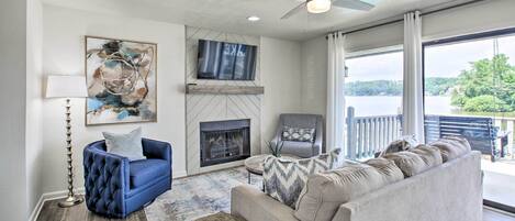 A living room with a fireplace, a ceiling fan, and a large window overlooking a lake.