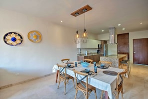 Un comedor con una mesa preparada para cuatro personas, rodeada de sillas de madera. Se ve una cocina al fondo con una encimera y armarios.