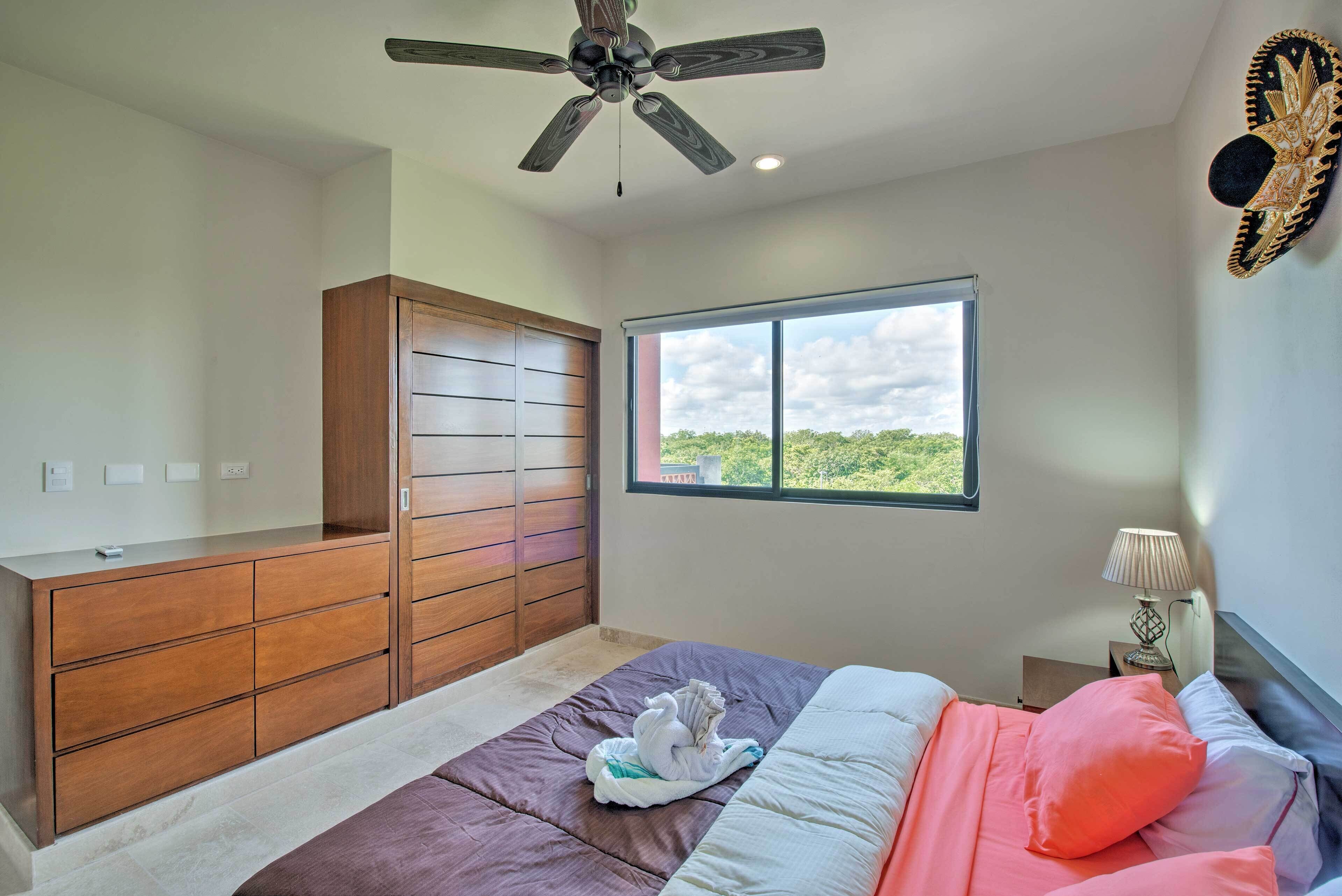 A bedroom with a bed, a wooden wardrobe, a ceiling fan, a window with a view, and a nightstand with a lamp.