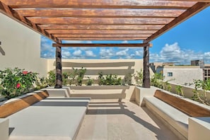 Un patio exterior con pérgola de madera, cojines color beige y plantas en macetas.